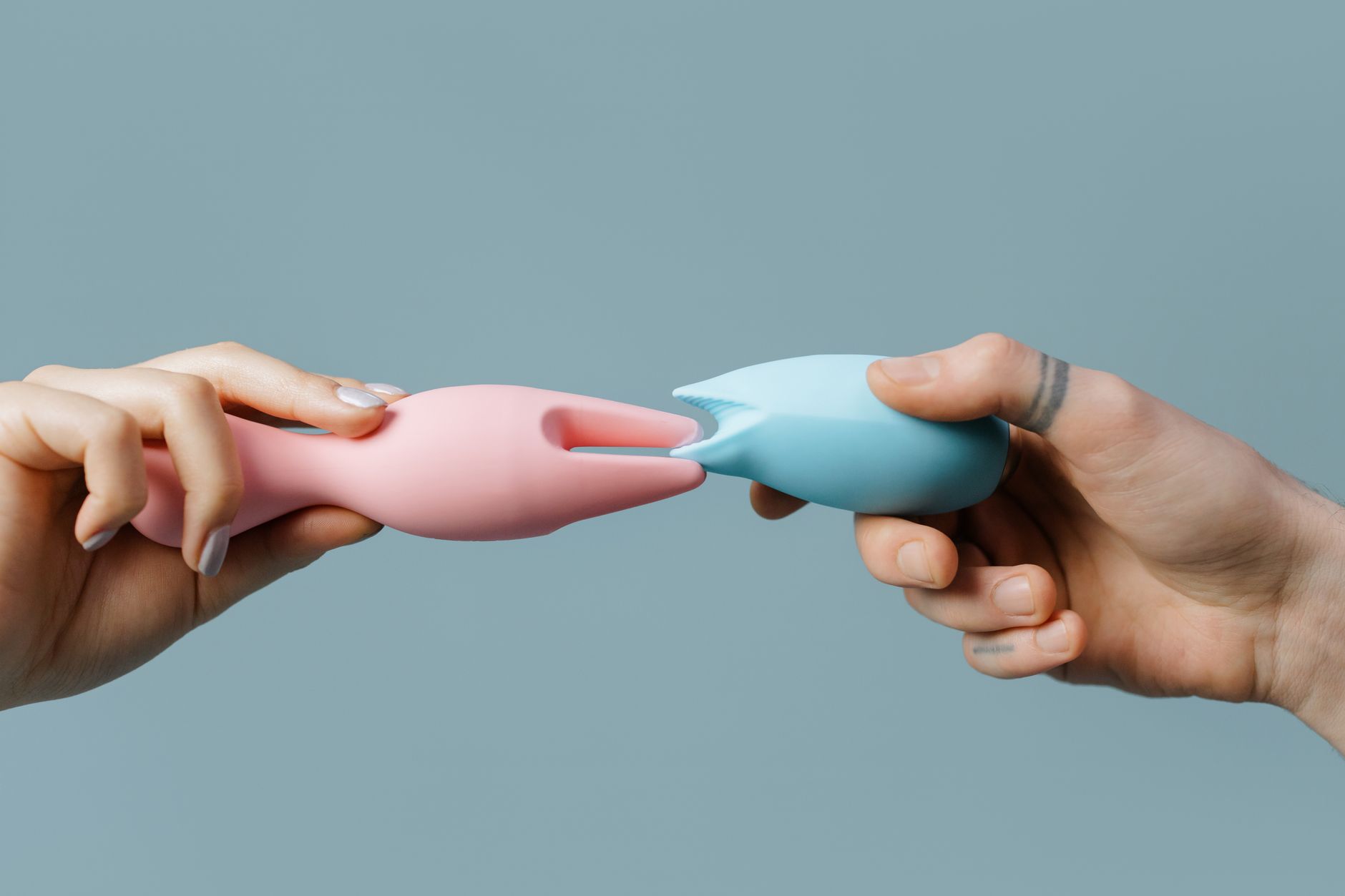 Two hands holding silicone vibrators against a pastel background, representing couple's shared pleasure and communication.